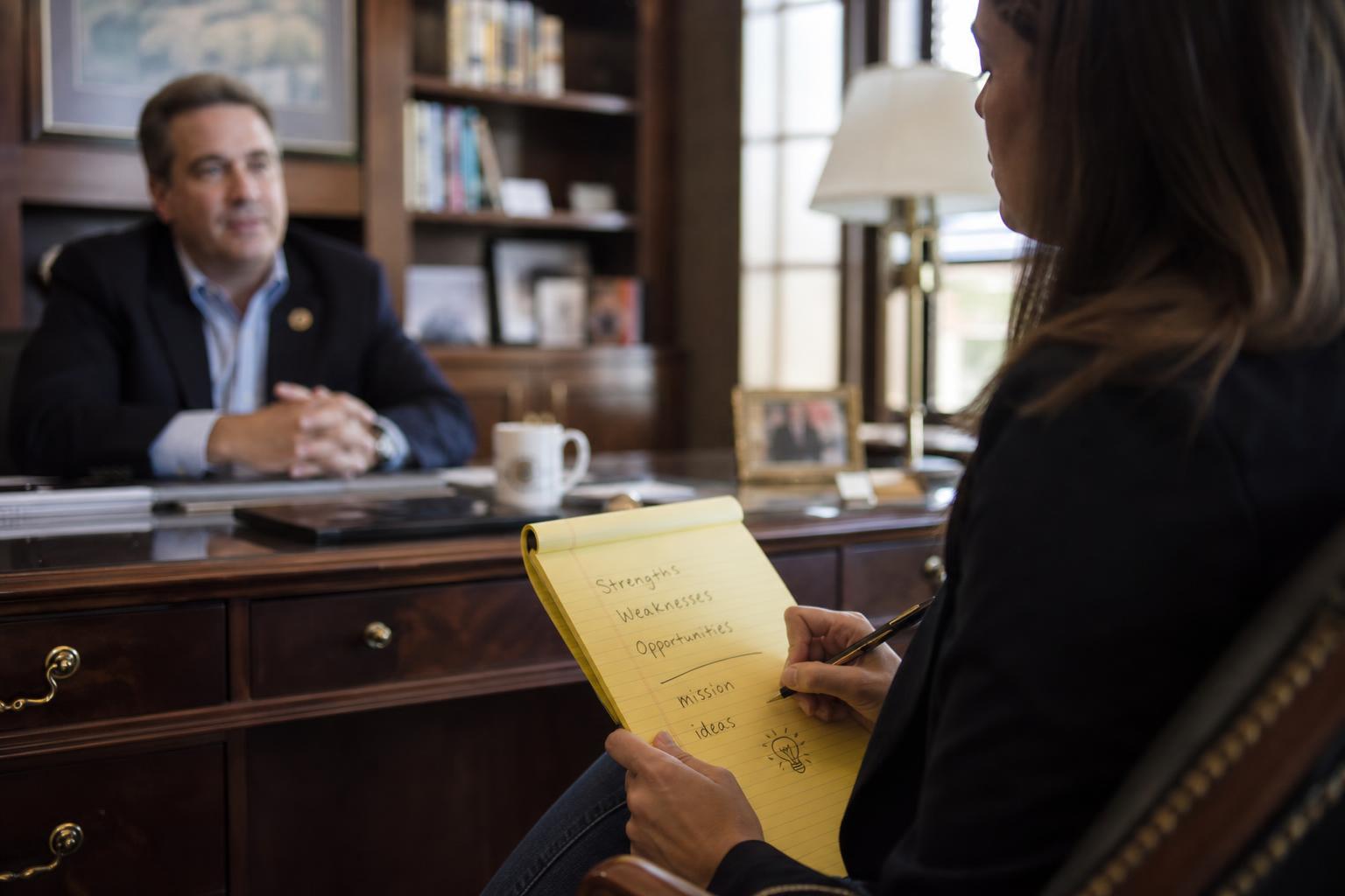 A consultant in profile view takes handwritten notes on a yellow legal pad while a leader speaks across a wooden desk in a traditional office